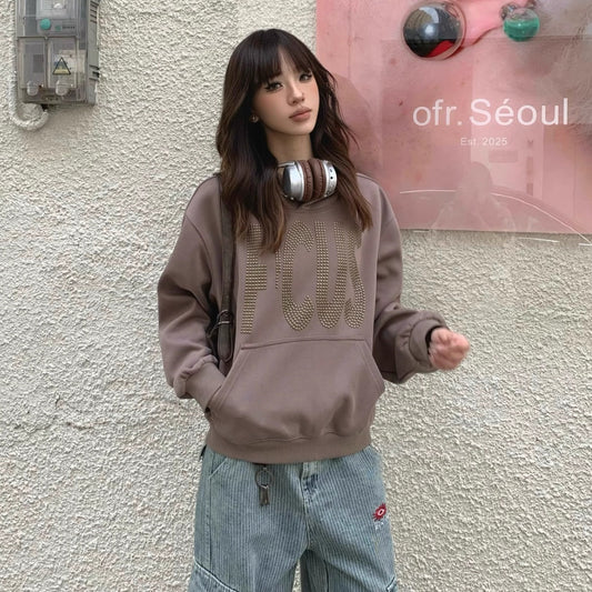 Model wearing oversized brown 'FOCUS' sweatshirt with studded lettering, styled with relaxed fit jeans and headphones around the neck, standing in front of a textured wall with pink Seoul art poster.