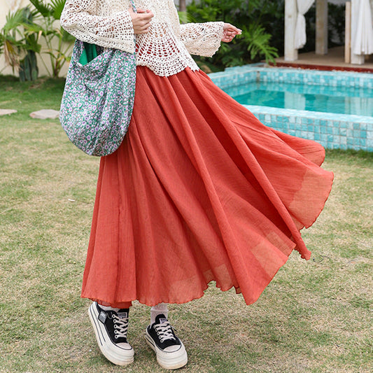 Flowy rust-orange maxi skirt styled with a white crochet top, floral shoulder bag, and chunky black sneakers, captured in an outdoor garden setting near a turquoise-tiled pool
