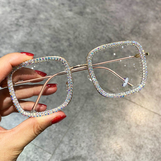 A pair of square-shaped, multicolored diamond-studded glasses with a metallic frame being held by a person with red nail polish.