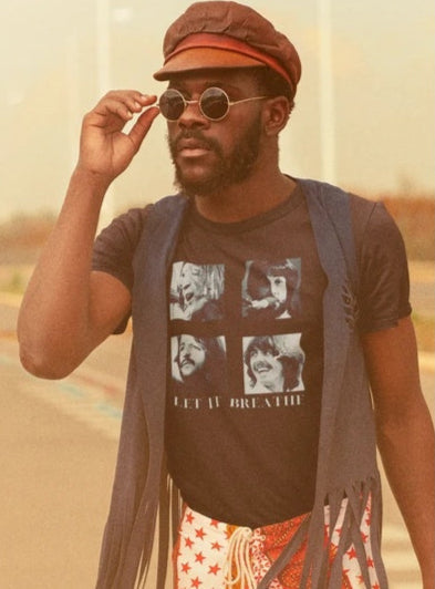 A person wearing a round-neck graphic t-shirt with the text 'LET IT BREATHE' and images of faces, paired with a fringed waistcoat and patterned trousers, accessorized with sunglasses and a hat.