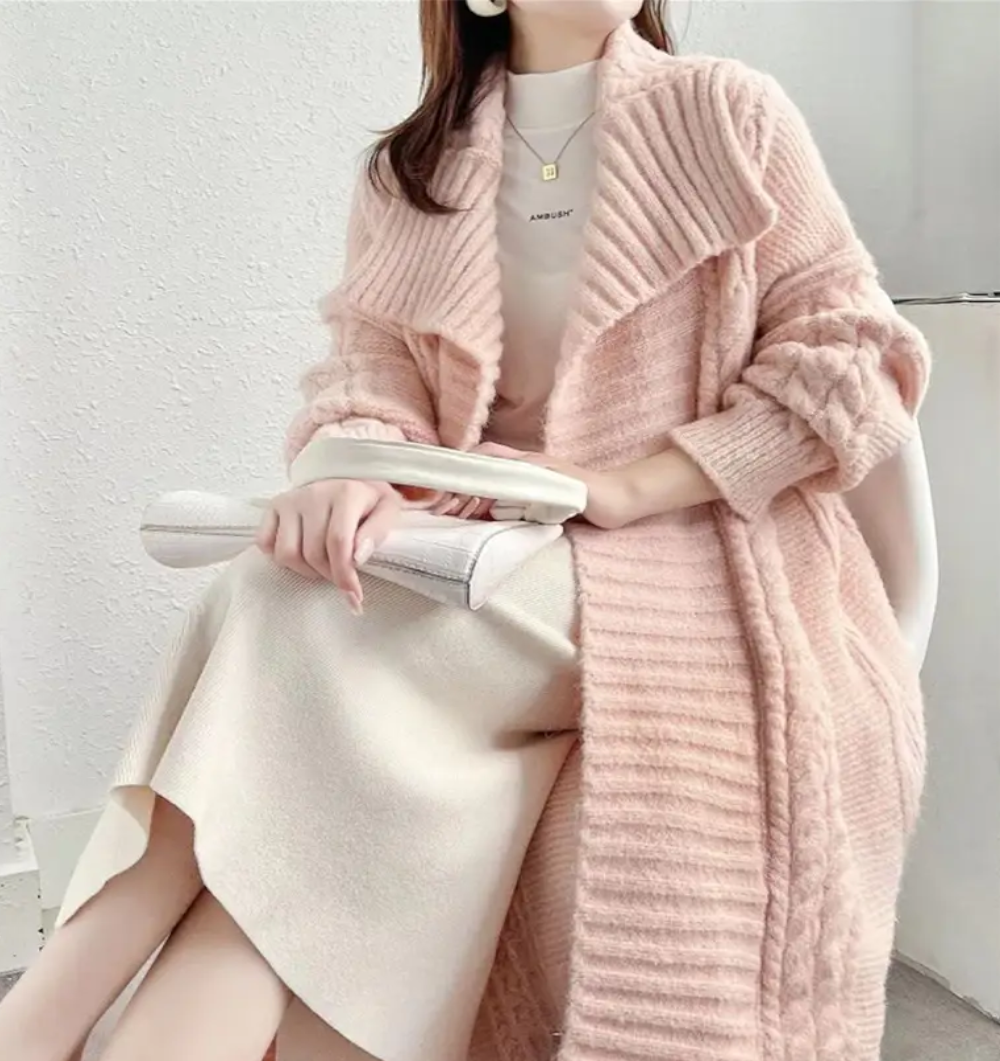 Person wearing a pink knitted cardigan over a white outfit, holding a white clutch.