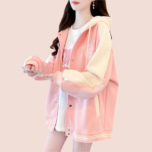 A woman wearing a pink and white long-sleeved baseball sweater with contrast color design and cartoon embroidery, holding a phone and standing indoors.