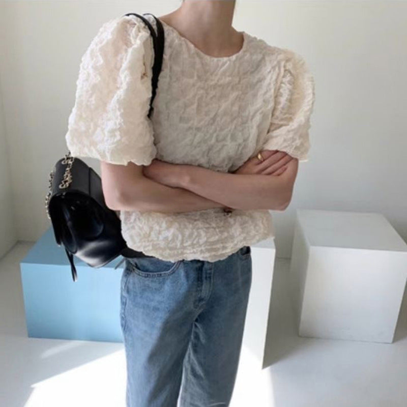 Person wearing a textured white top and blue jeans, holding a black bag against a minimal background.