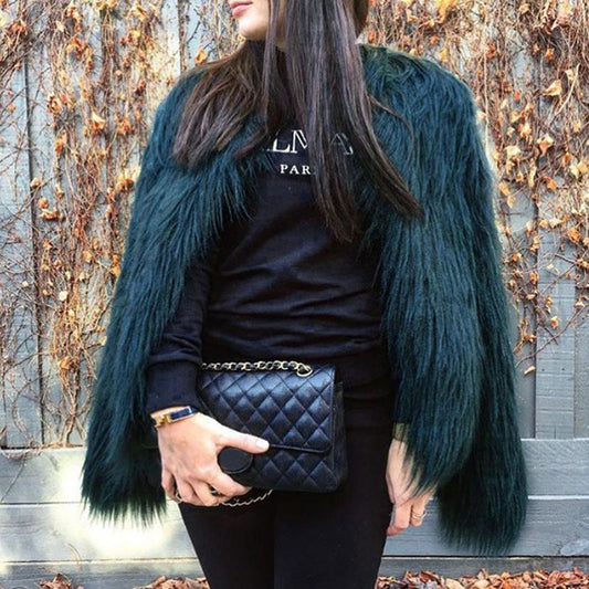 Person wearing a dark blue fur coat over a black outfit with a quilted handbag, standing against a leafless tree background.