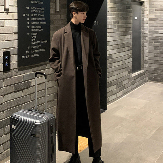 Man in a long coat standing next to a suitcase in a hallway with a brick wall.