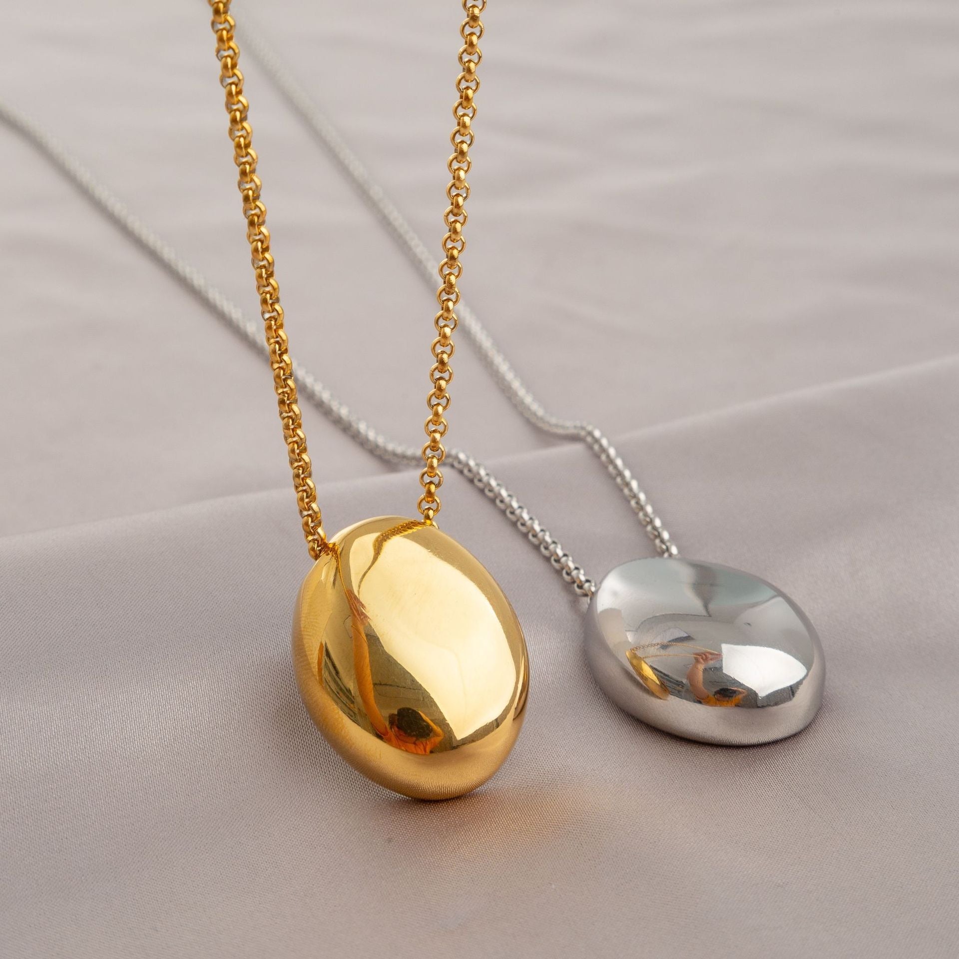 Minimalist oval pendant necklaces in polished gold and silver finishes, displayed on soft satin fabric for an elegant and modern look.