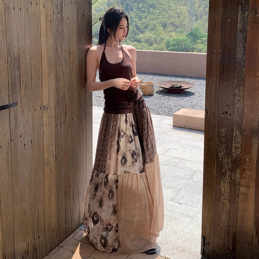 Woman in a long, patterned dress standing in a sunlit outdoor area with wooden walls and greenery.