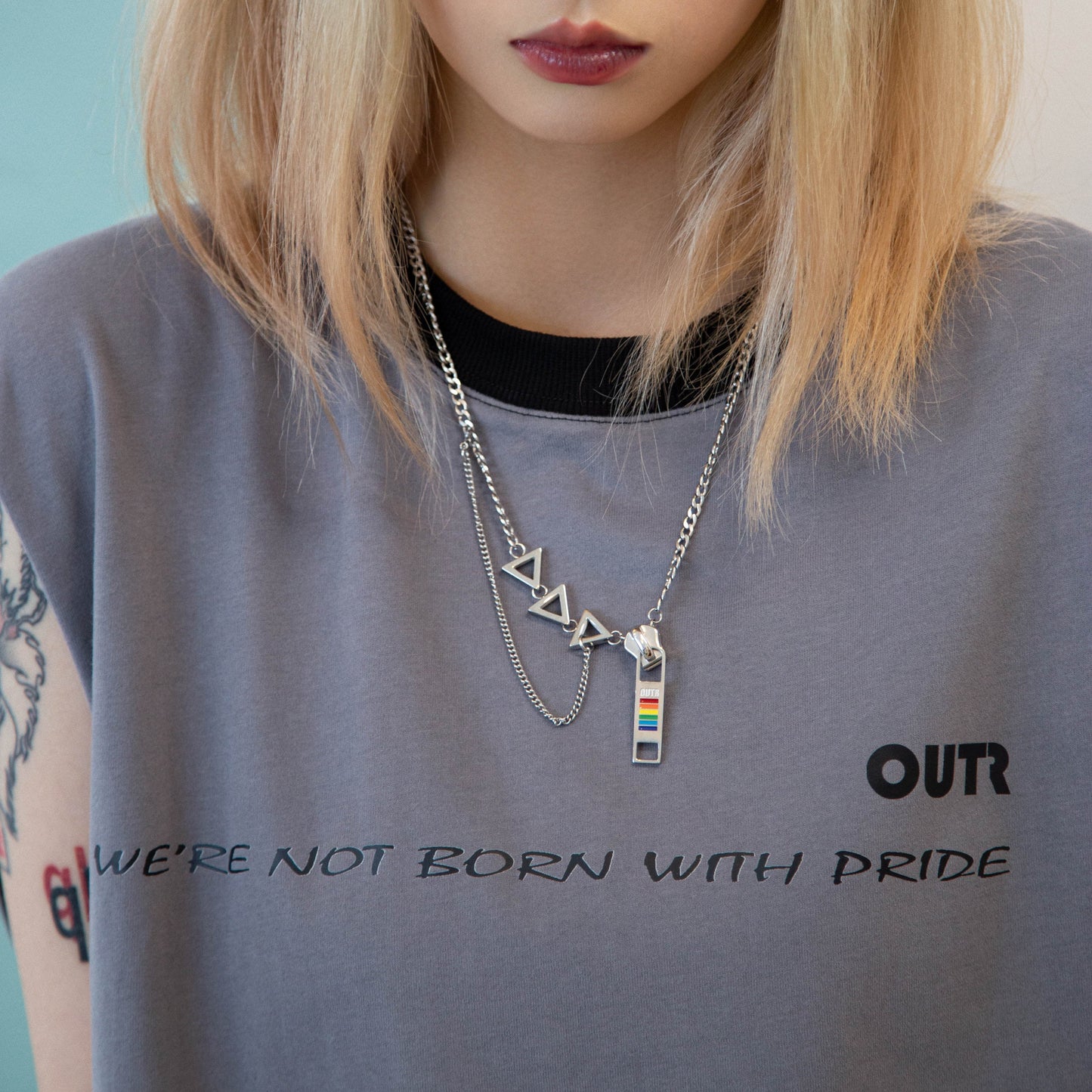 Person wearing a gray t-shirt with 'OUTR' and 'We're Not Born with Pride' text, and multiple necklaces.