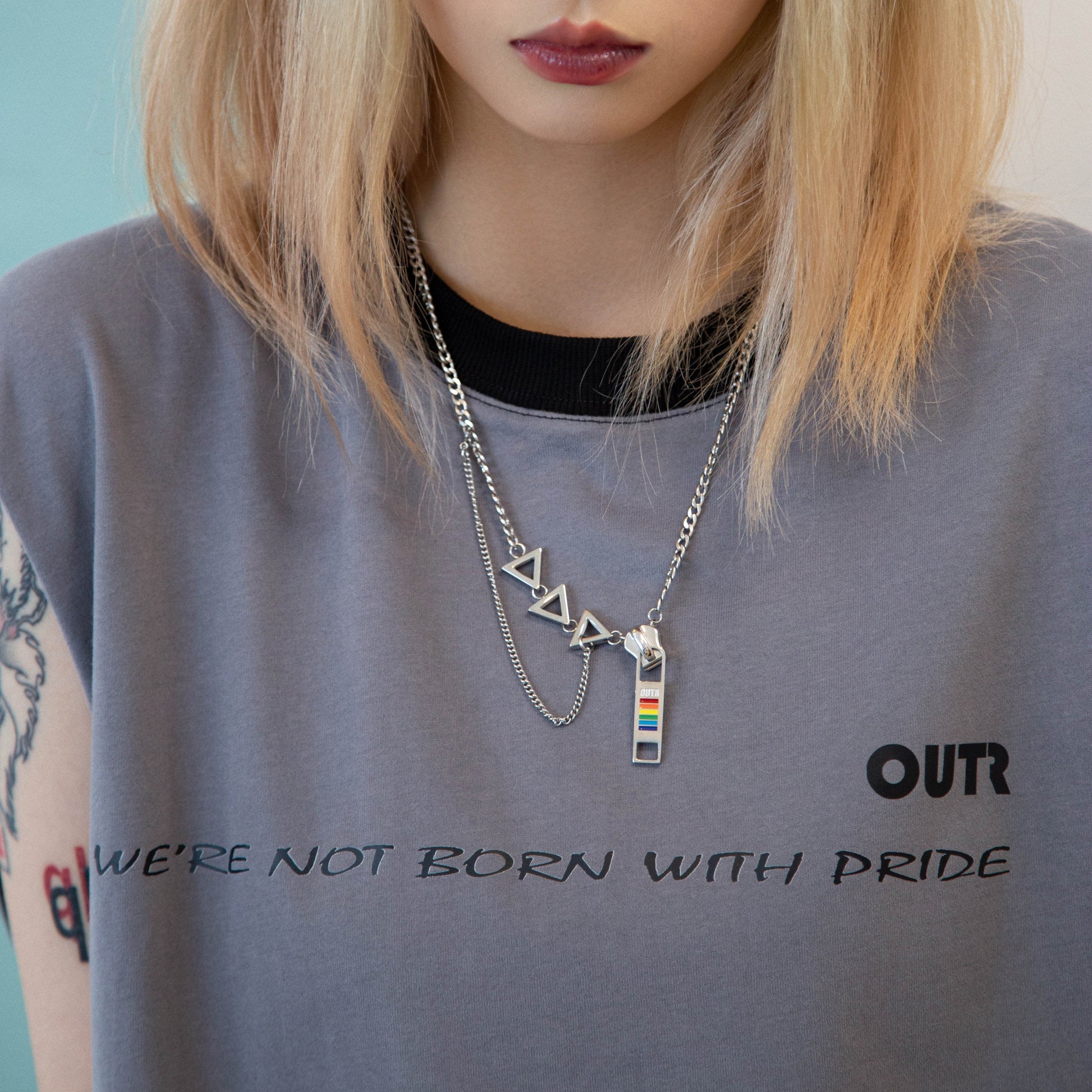 Person wearing a gray t-shirt with 'OUTR' and 'We're Not Born with Pride' text, and multiple necklaces.