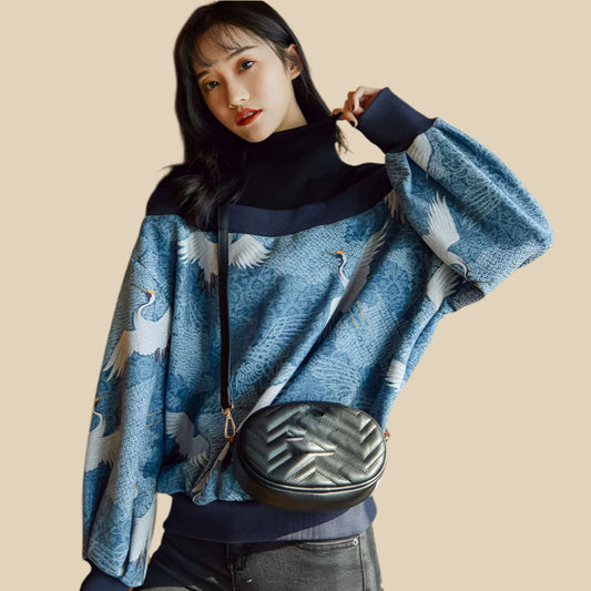 A woman wearing a blue crane print casual sweater with a high neck, lantern sleeves, and black pants. She is carrying a black bag and the setting appears to be indoors with wooden panels in the background.