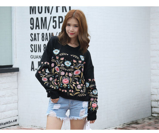 Woman wearing a black floral embroidered sweater and denim shorts standing against a white wall.