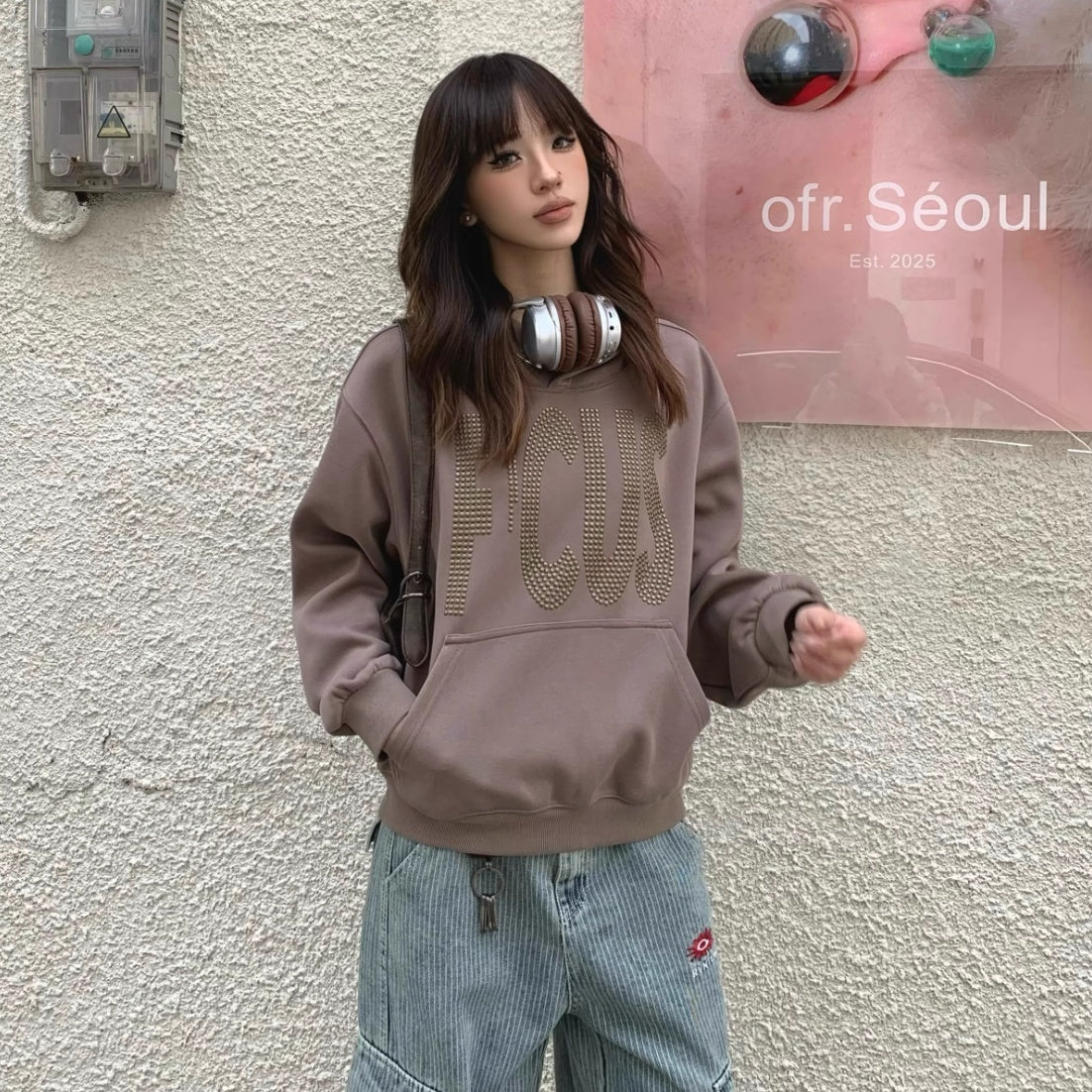 Model wearing oversized brown 'FOCUS' sweatshirt with studded lettering, styled with relaxed fit jeans and headphones around the neck, standing in front of a textured wall with pink Seoul art poster.