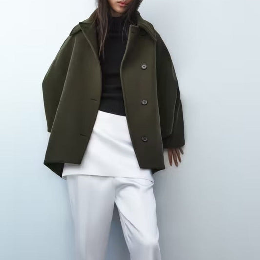 Person wearing an olive green coat over a black top and white pants on a light blue background