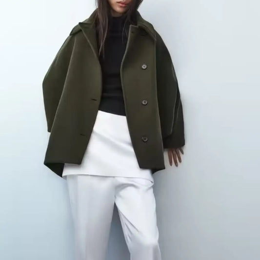 Person wearing an olive green coat over a black top and white pants on a light blue background