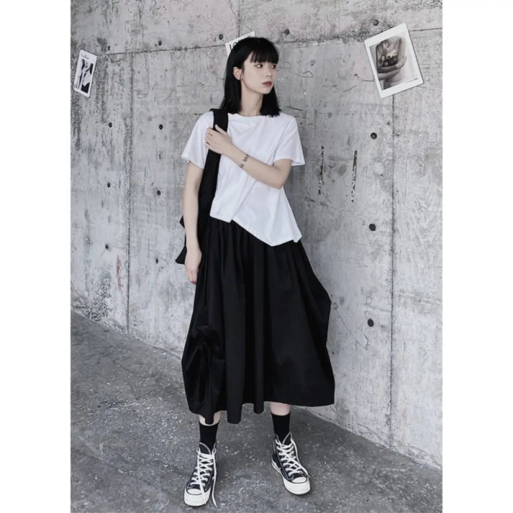 Person wearing a white t-shirt and black skirt against a concrete wall.