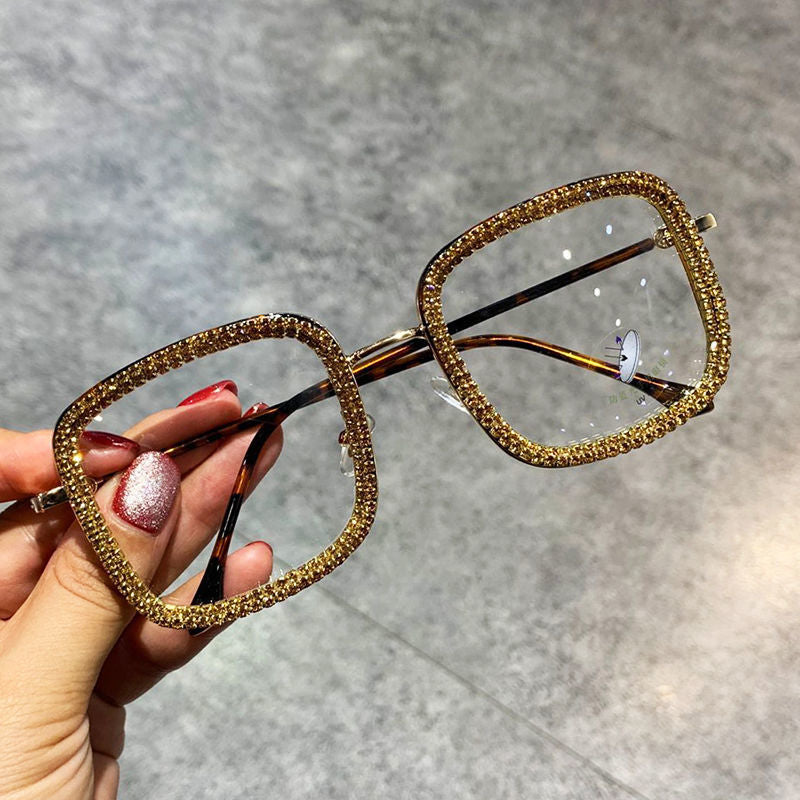 A pair of square-shaped, multicolored diamond-studded glasses with a metallic frame being held by a person with red nail polish.