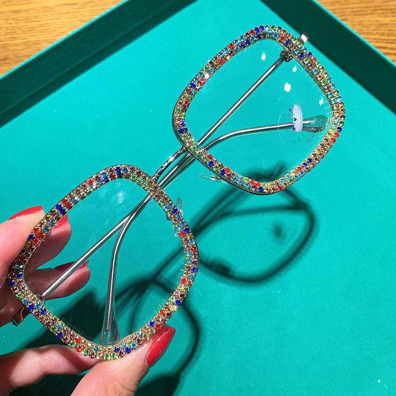 A pair of square-shaped, multicolored diamond-studded glasses with a metallic frame being held by a person with red nail polish.