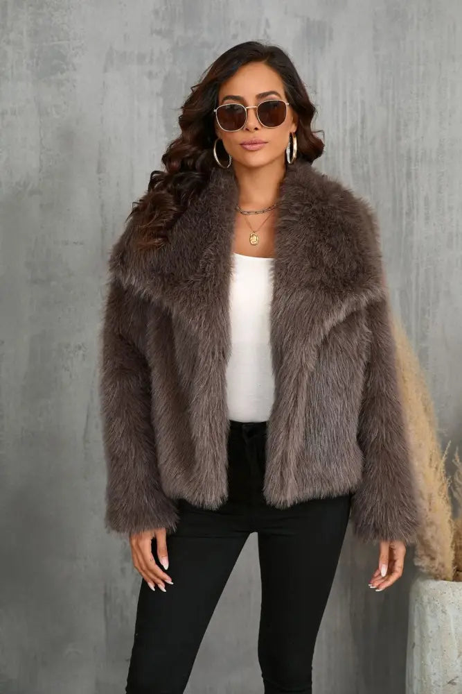 A female model wearing a large lapel fluffy plush artificial fur coat in brown color with long sleeves.