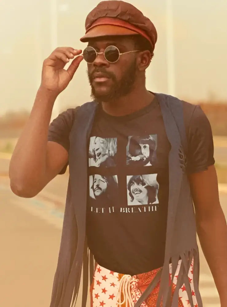 A person wearing a round-neck graphic t-shirt with the text 'LET IT BREATHE' and images of faces, paired with a fringed waistcoat and patterned trousers, accessorized with sunglasses and a hat.