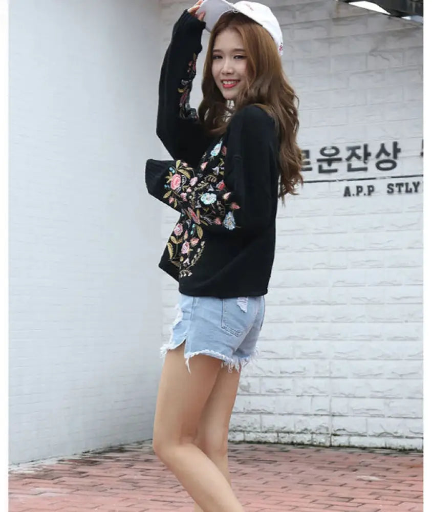 Woman wearing a black floral hoodie and denim shorts against a white wall.