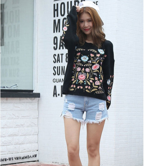 Woman wearing a black floral embroidered sweater and denim shorts standing in front of a white wall with text.