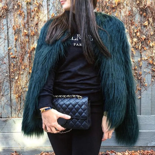 Person wearing a dark blue fur coat over a black outfit with a quilted handbag, standing against a leafless tree background.