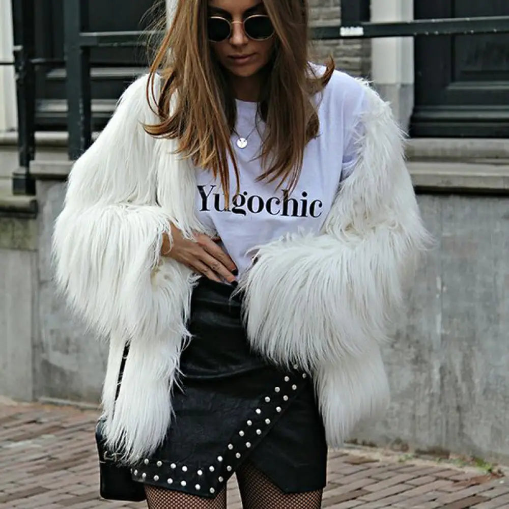 Person wearing a white fur coat, white t-shirt with text, and black skirt with studs outdoors.