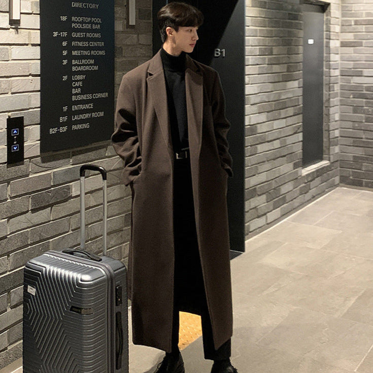 Man in a long coat standing next to a suitcase in a hallway with a brick wall.