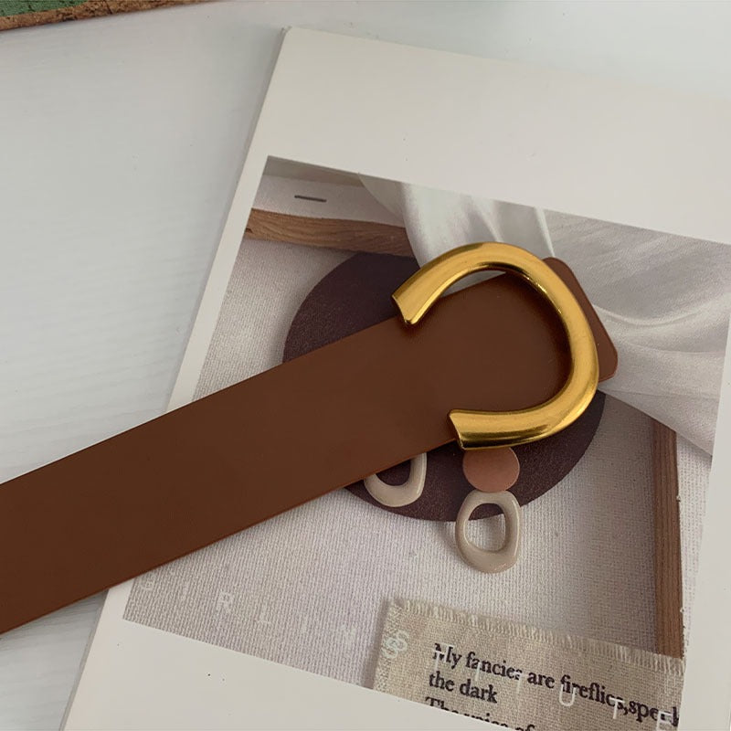 A brown belt with a gold-colored buckle placed on a flat surface, partially lying on a white object which appears to be a piece of paper or fabric.